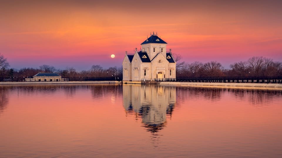 A historic building with a tower, reflecting in a calm body of water, with a sunset sky in the background.