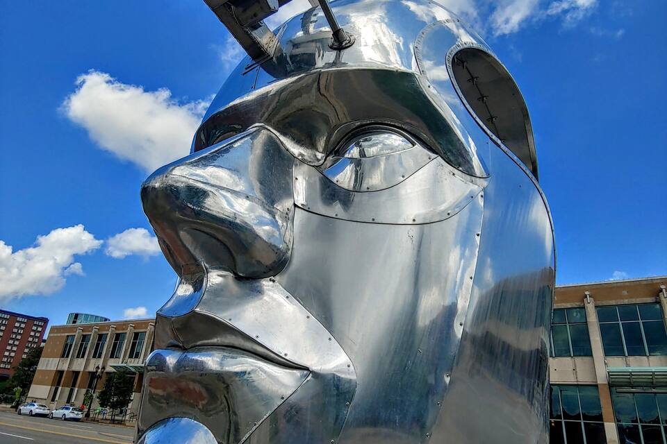 A large metallic sculpture of a face with a helmet, set against a clear blue sky with clouds.