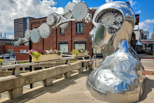 Une grande sculpture métallique représentant une tête ornée d’engrenages, placée devant un bâtiment en briques et un ciel bleu dégagé.
