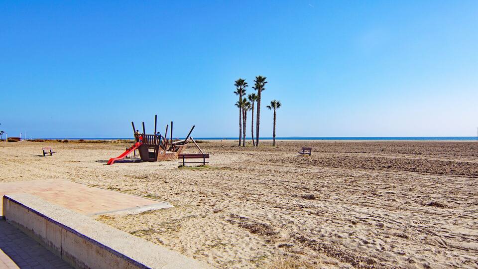 A sandy beach with palm trees, a red slide, and benches.