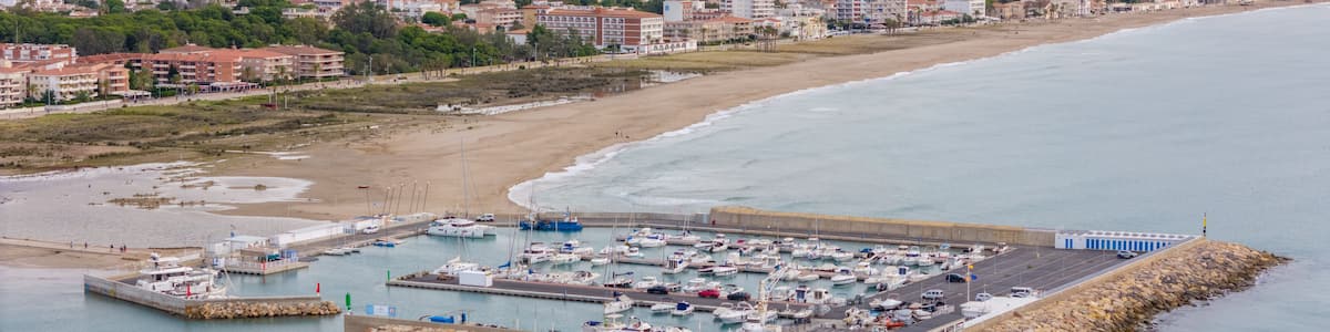 A coastal marina with boats docked, a sandy beach, and a cityscape in the background.