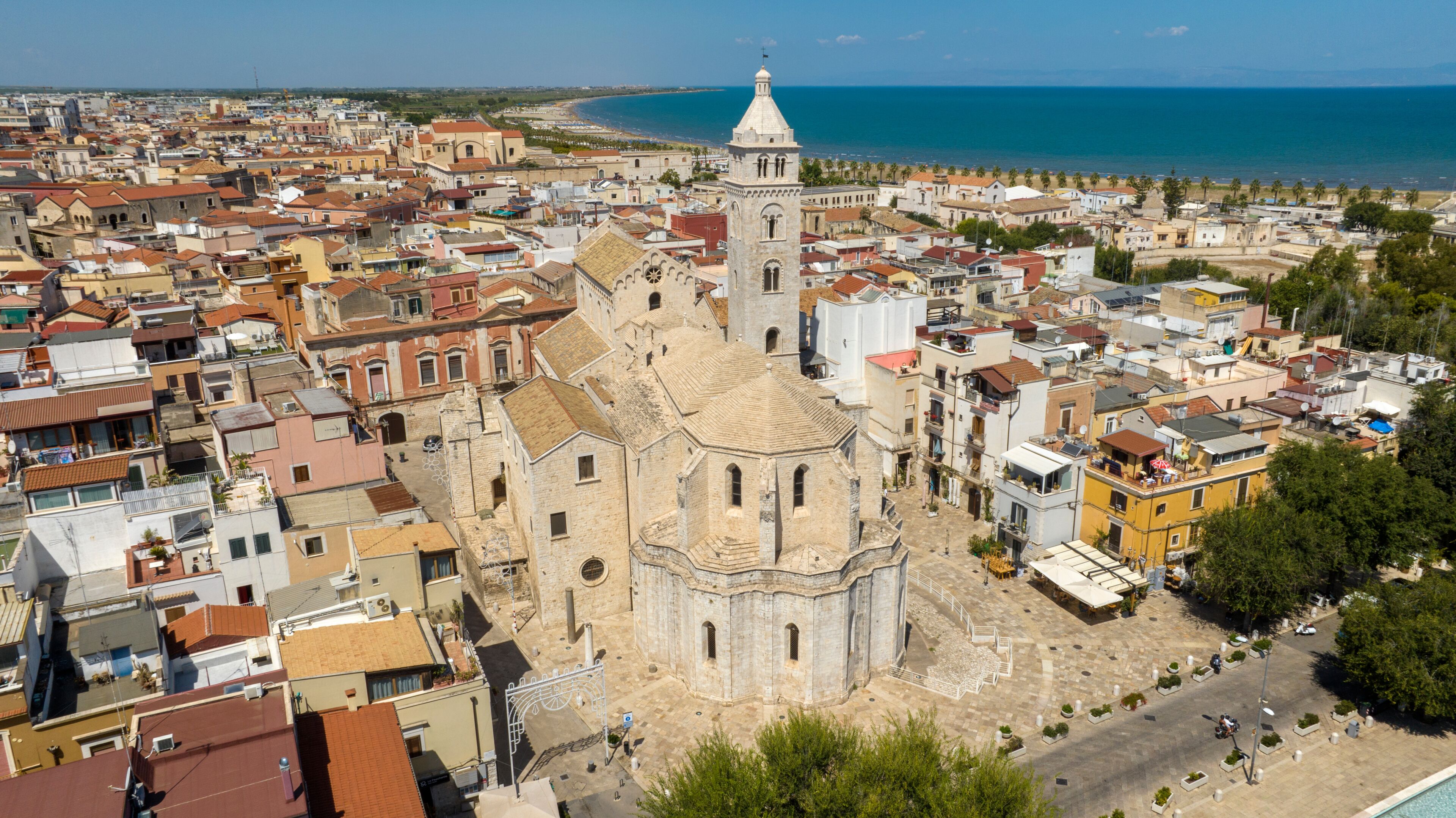 A coastal cityscape with a prominent church featuring a bell tower, surrounded by dense urban buildings and a beach in the background.