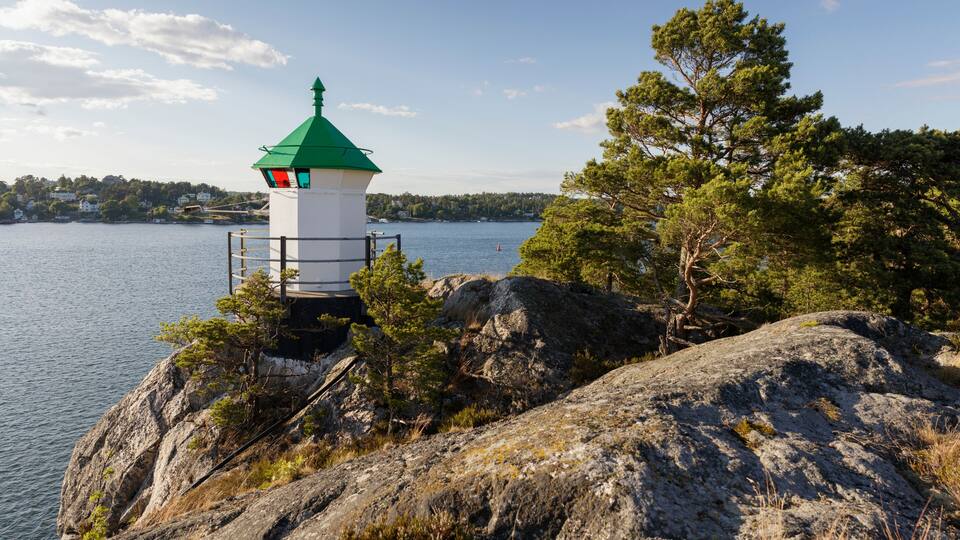 A lighthouse on a rocky outcrop by a lake with a green roof, surrounded by trees and a clear sky.