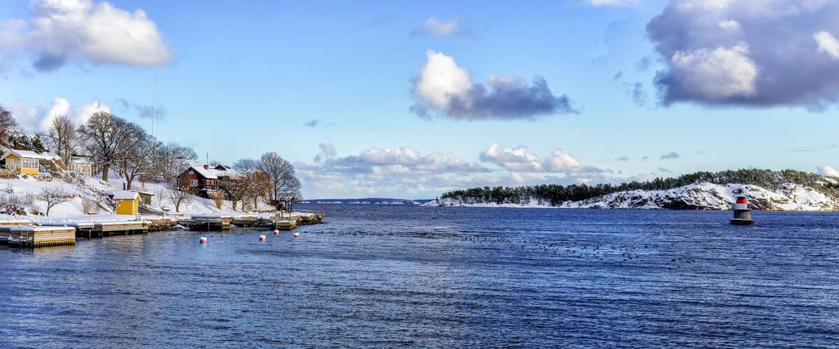 A snowy waterfront with docks, a lighthouse, and a snow-covered island in the distance.
