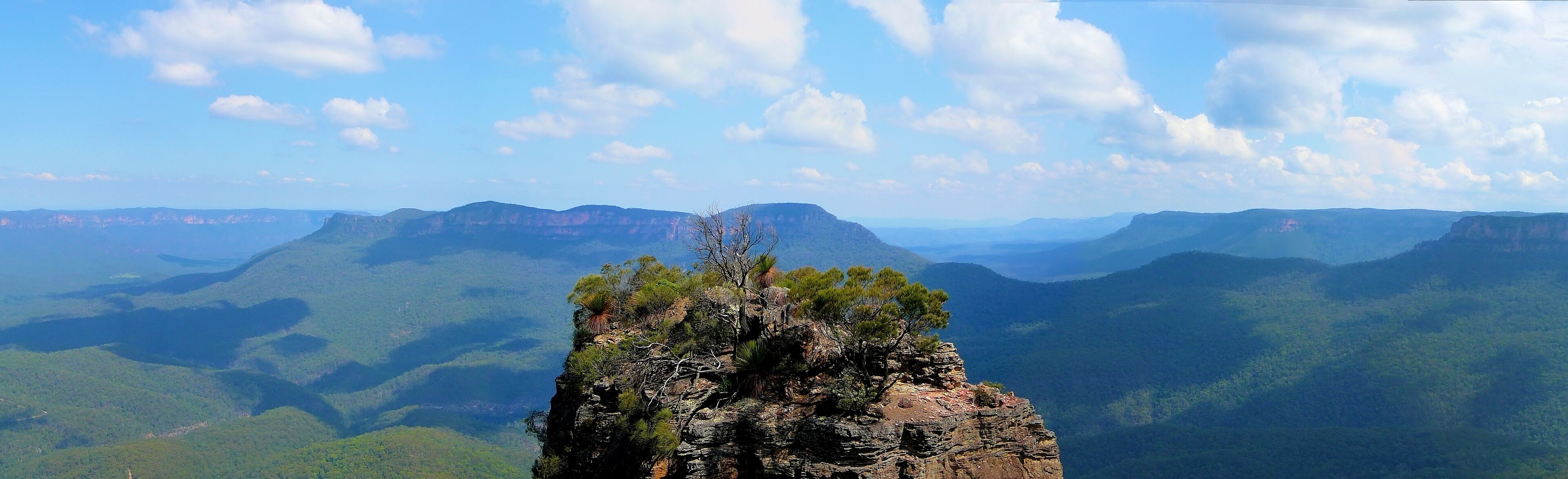 A rugged mountain peak with sparse vegetation, surrounded by expansive green valleys and distant mountain ranges under a clear sky.