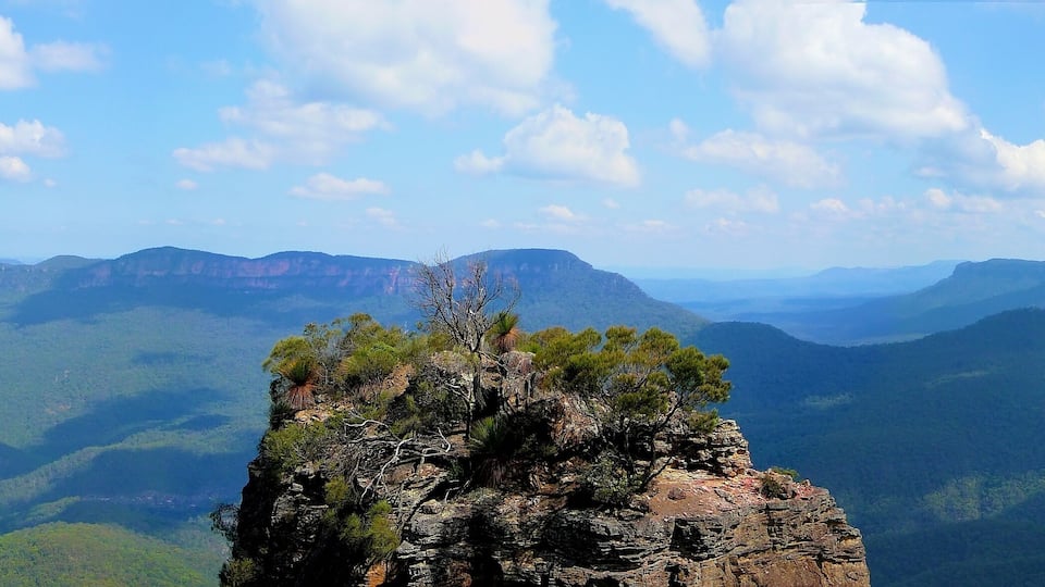 A rugged mountain peak with sparse vegetation, surrounded by expansive green valleys and distant mountain ranges under a clear sky.