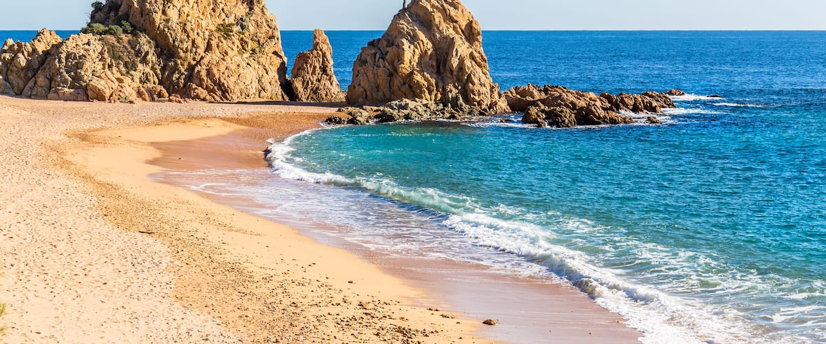 A sandy beach with rocky formations and a clear blue sea.