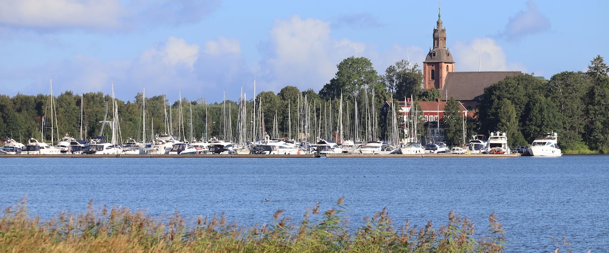 A harbor with numerous sailboats and yachts docked, a prominent church with a tall steeple, and a backdrop of trees and a clear sky.