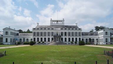 A large, symmetrical building with a central entrance, surrounded by a well-manicured lawn and rows of pink flowers.