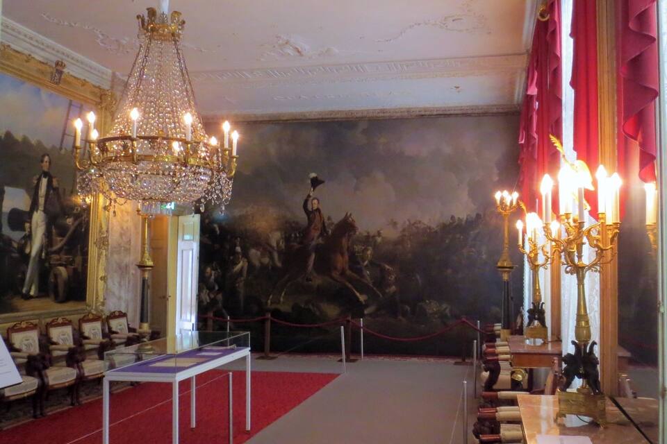 An ornate room with a large chandelier, a red carpet, and a historical painting on the wall.