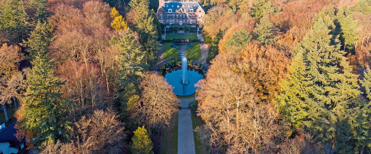 An aerial view of a large estate surrounded by dense forest, with a prominent building featuring a tower and a symmetrical driveway.