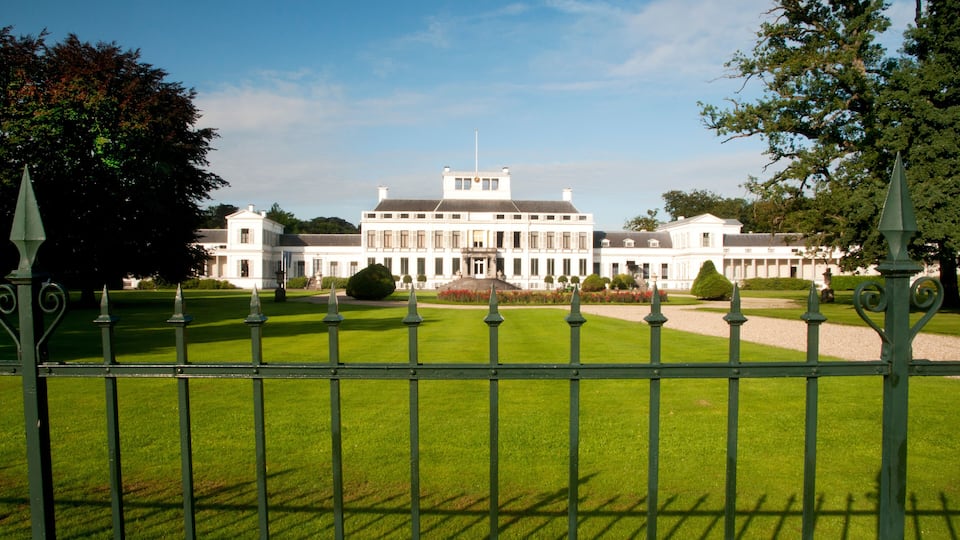 A large white building with a symmetrical facade, surrounded by a well-manicured lawn and a green metal fence.