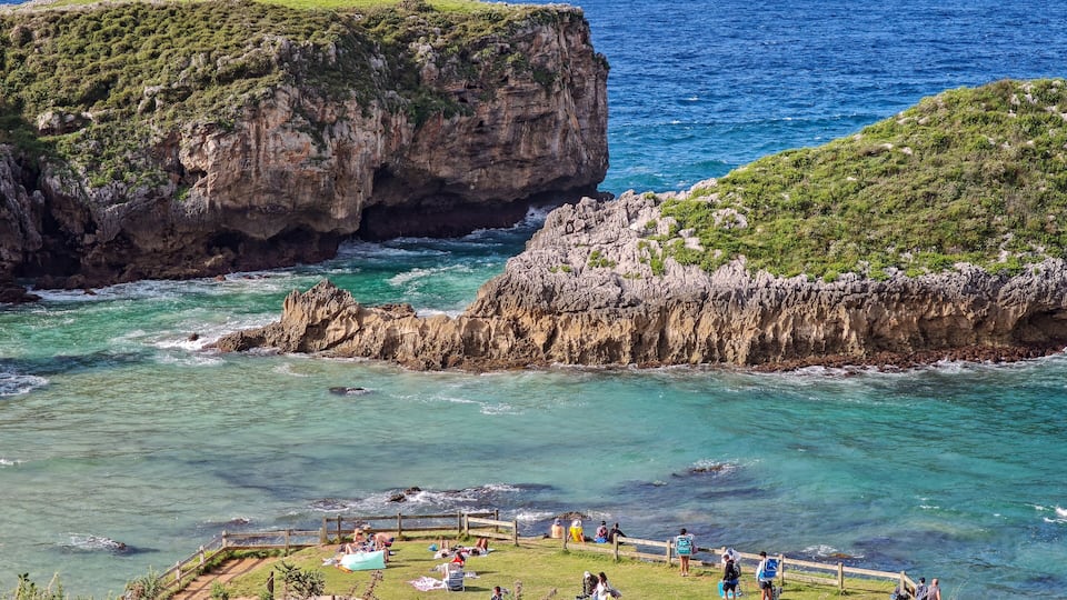 A coastal landscape with cliffs, clear turquoise waters, and people relaxing on the beach.