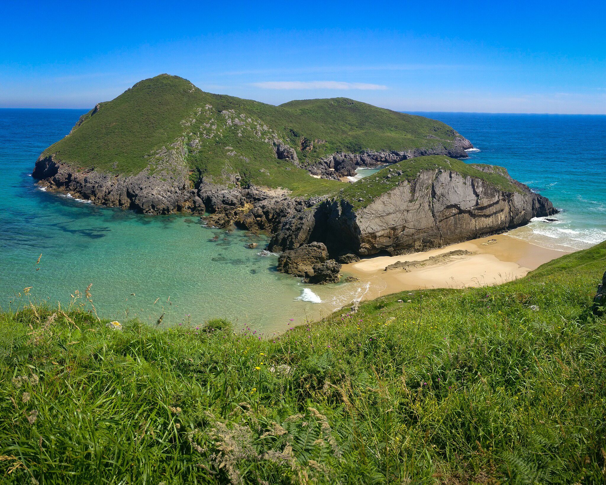 A coastal landscape with green hills, clear turquoise waters, and a sandy beach.