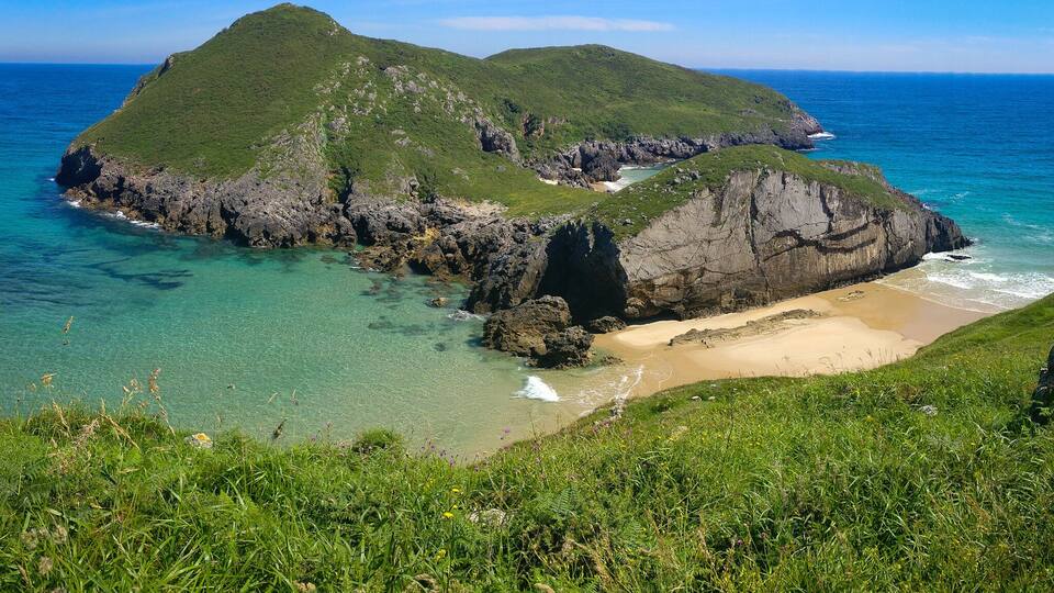 A coastal landscape with green hills, clear turquoise waters, and a sandy beach.
