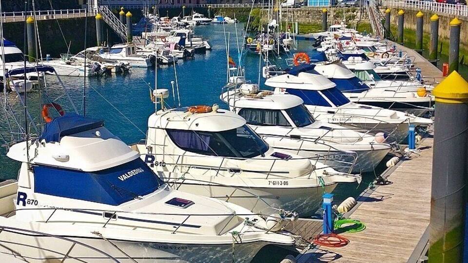 A marina with numerous boats docked along a wooden pier, surrounded by buildings and mountains in the background.