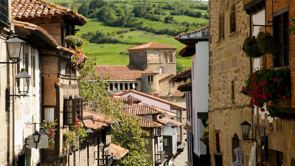 A narrow street with traditional buildings, a tower, and a green hillside in the background.