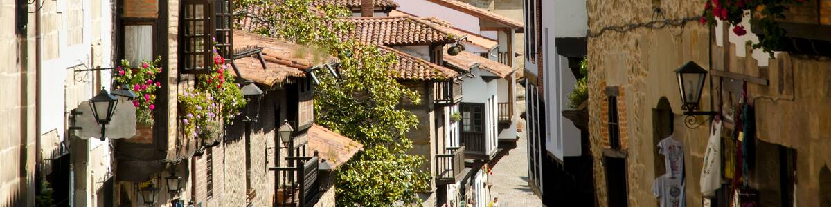 A narrow street with traditional buildings, a tower, and a green hillside in the background.