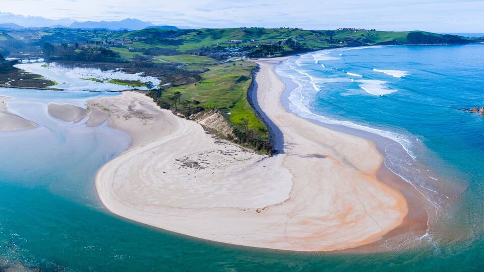 A coastal landscape with a sandy beach, a river mouth, and green hills in the background.