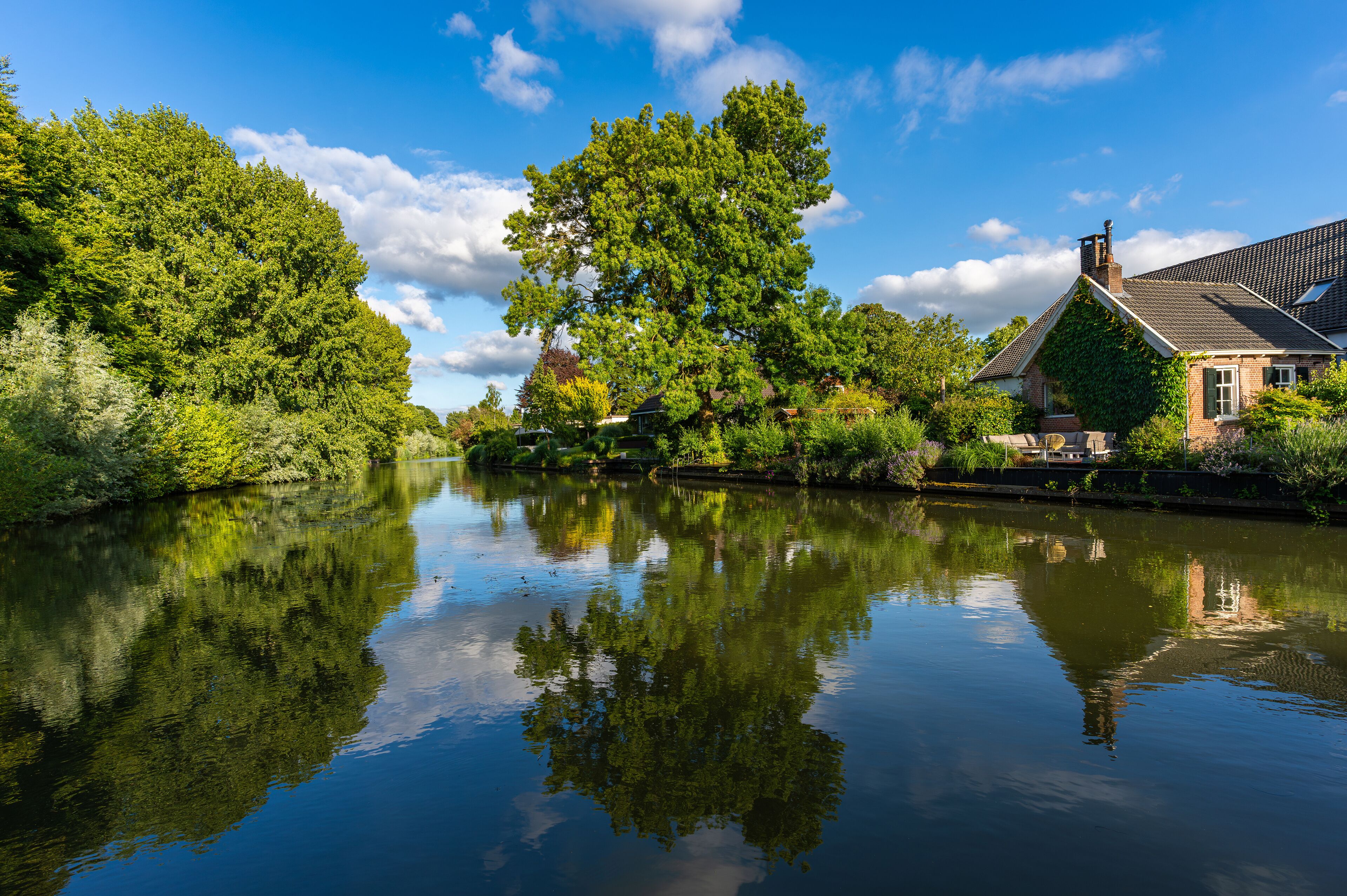 A calm river reflecting trees and a house with a sloped roof.