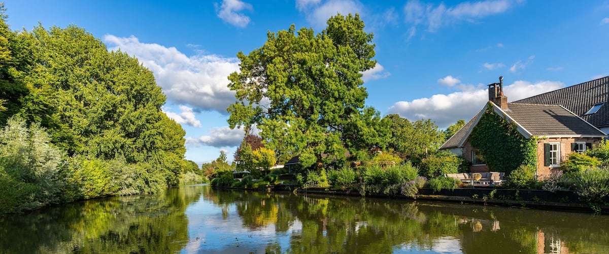 A calm river reflecting trees and a house with a sloped roof.