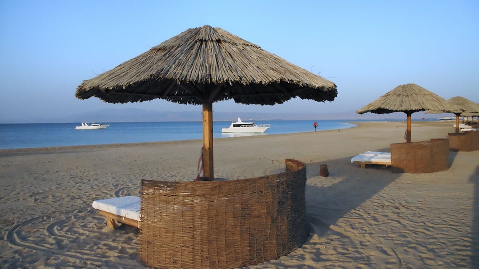 A beach with thatched umbrellas, lounge chairs, and a boat in the water.