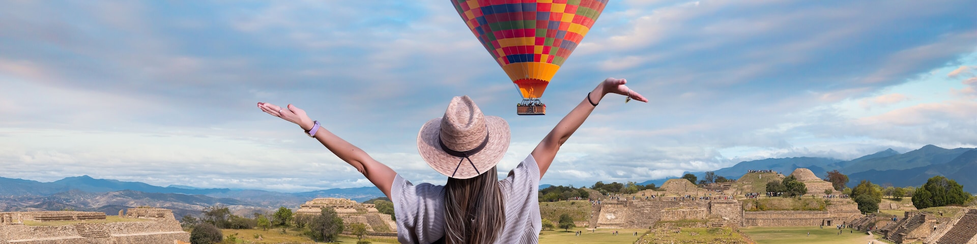 Une personne les bras levés, un ballon à air chaud aux couleurs vives, et des ruines antiques en arrière-plan.