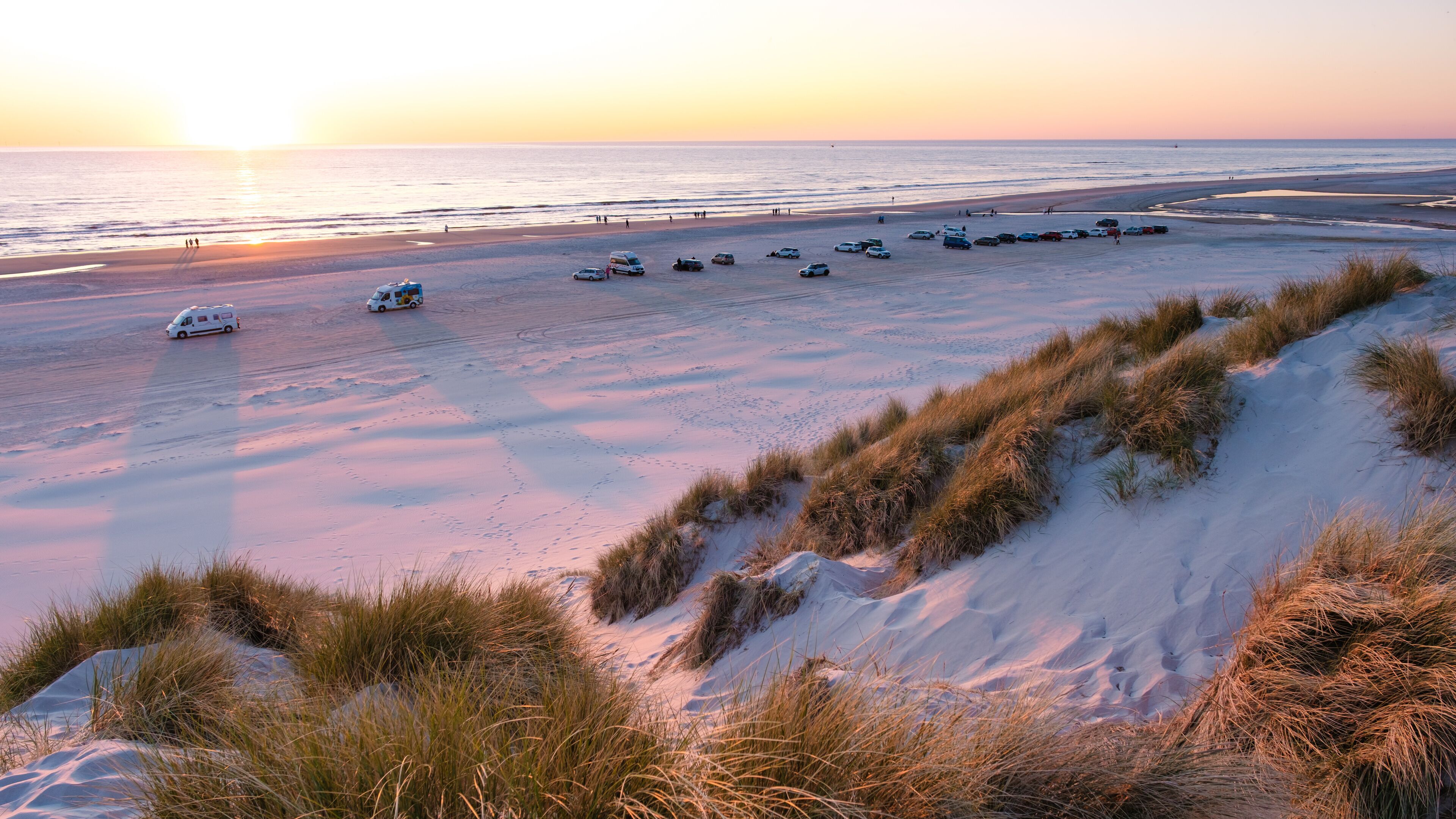 A beach at sunset with parked cars and dunes.