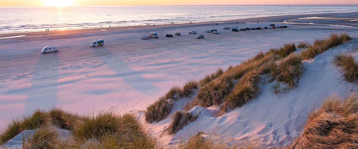 A beach at sunset with parked cars and dunes.