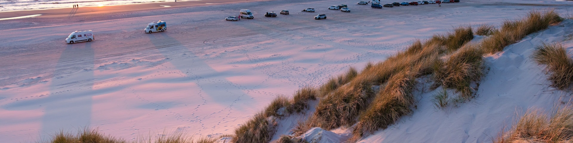 A beach at sunset with parked cars and dunes.
