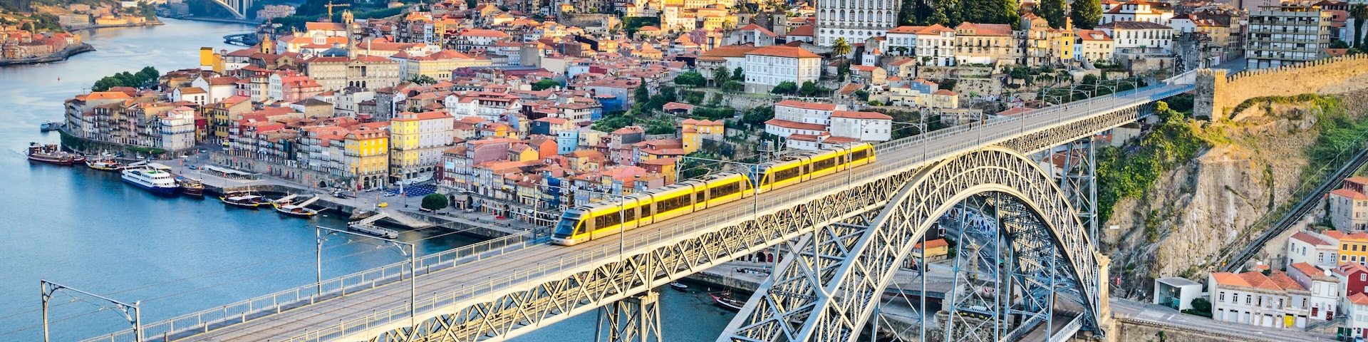 A yellow train on a steel bridge over a river, with historic buildings and a hill in the background.