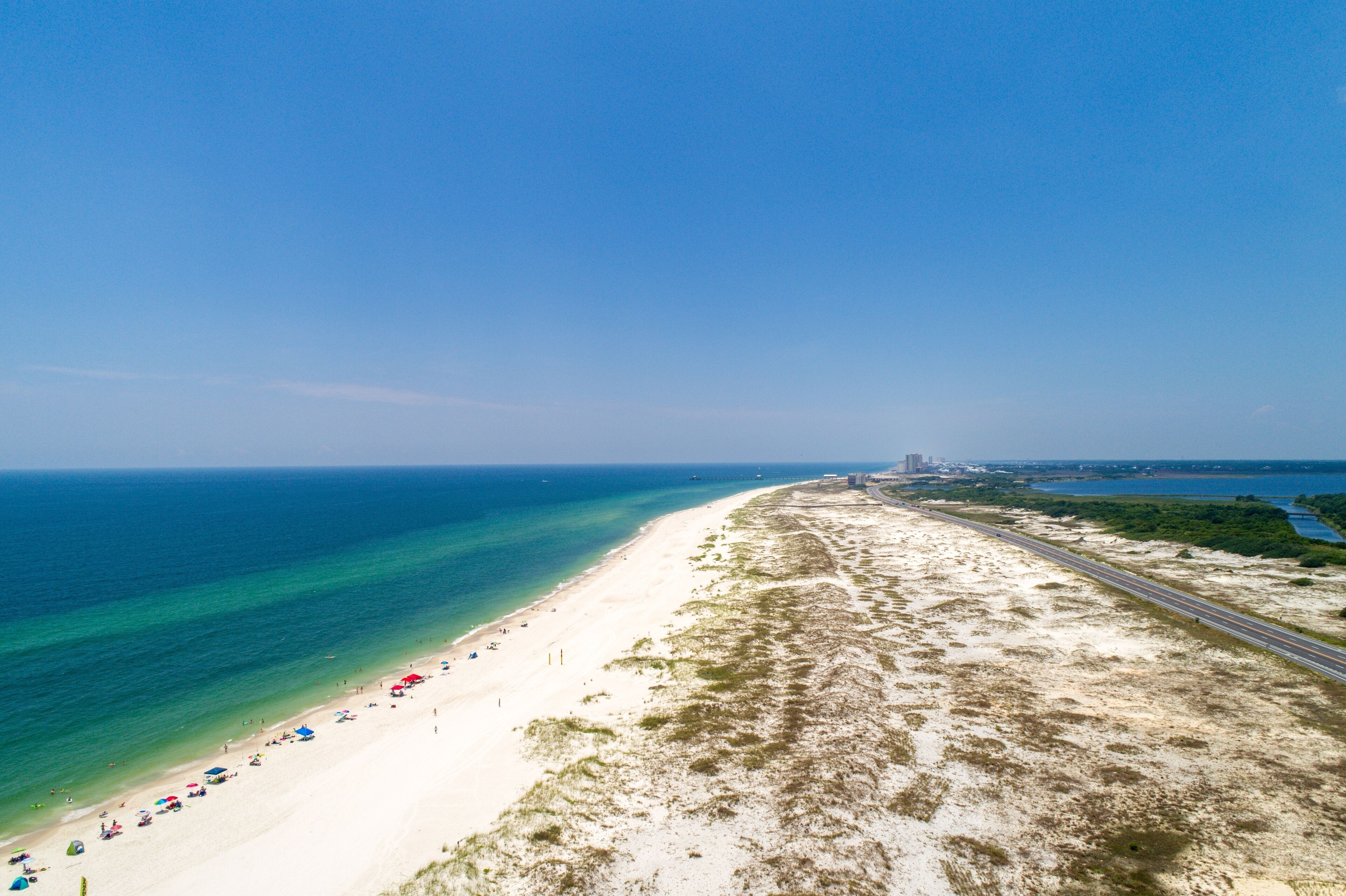 A coastal beach with clear blue waters, a sandy shore, and a highway running parallel to the beach.