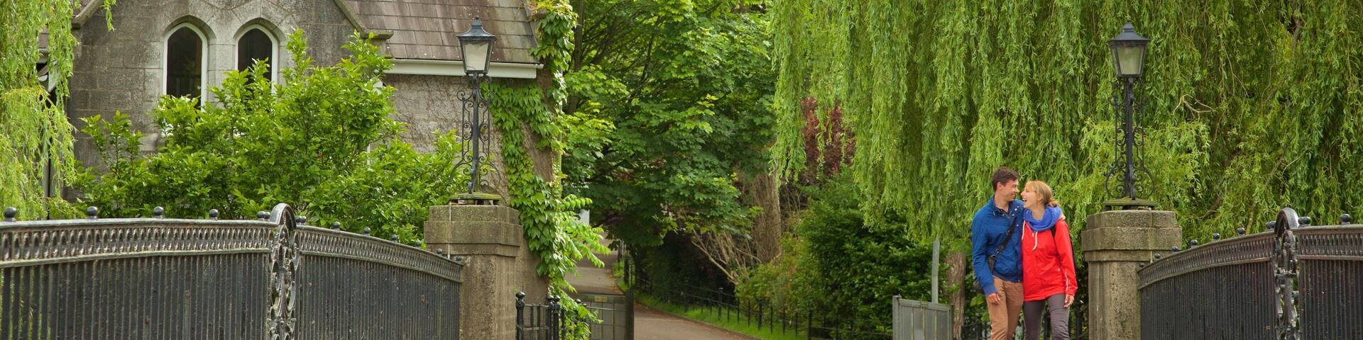 A stone bridge with ornate metal railings, a historic building with a pointed roof, and lush green trees.
