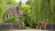 A stone bridge with ornate metal railings, a historic building with a pointed roof, and lush green trees.
