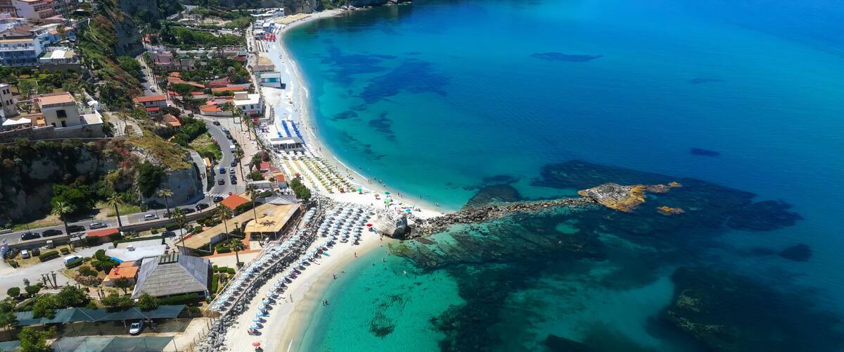 A coastal town with a long sandy beach, clear turquoise waters, and rocky outcrops.