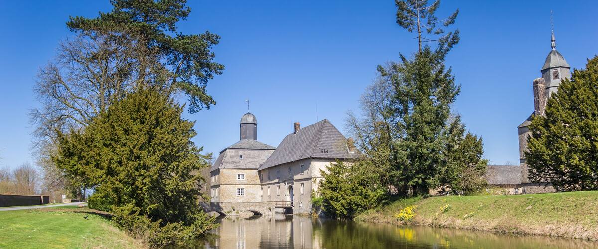 A historic building with a large tower, a bridge over a calm river, and surrounding trees.