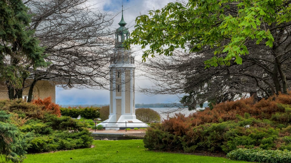 A white tower with a green spire, surrounded by lush greenery and a body of water in the background.