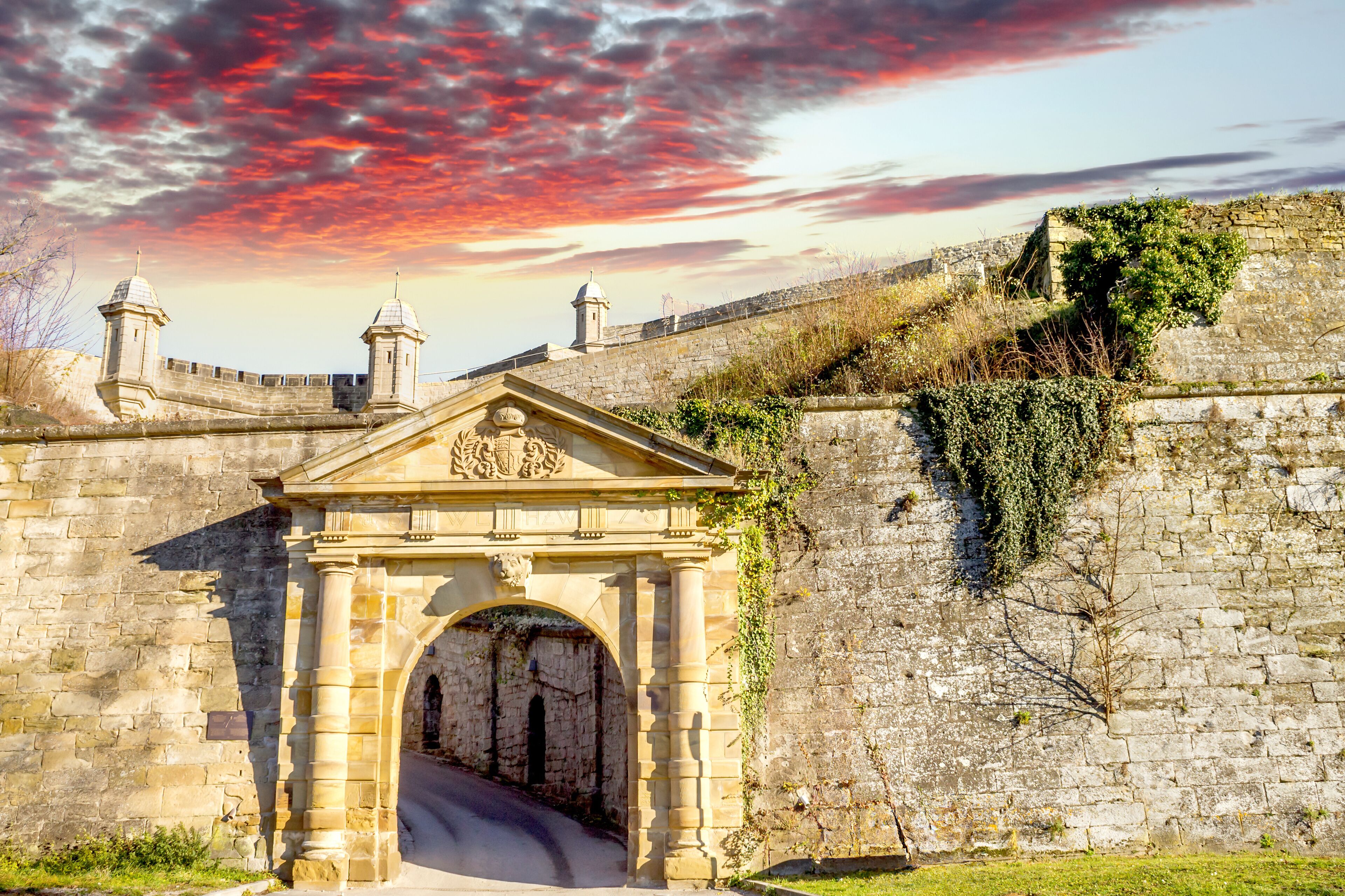 A stone archway with a decorative crest, a stone wall with vegetation, and a tower in the background.