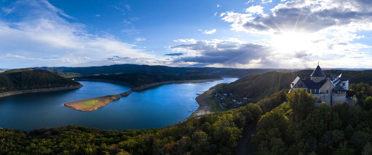A panoramic view of a lake surrounded by hills, a castle, and a clear sky.