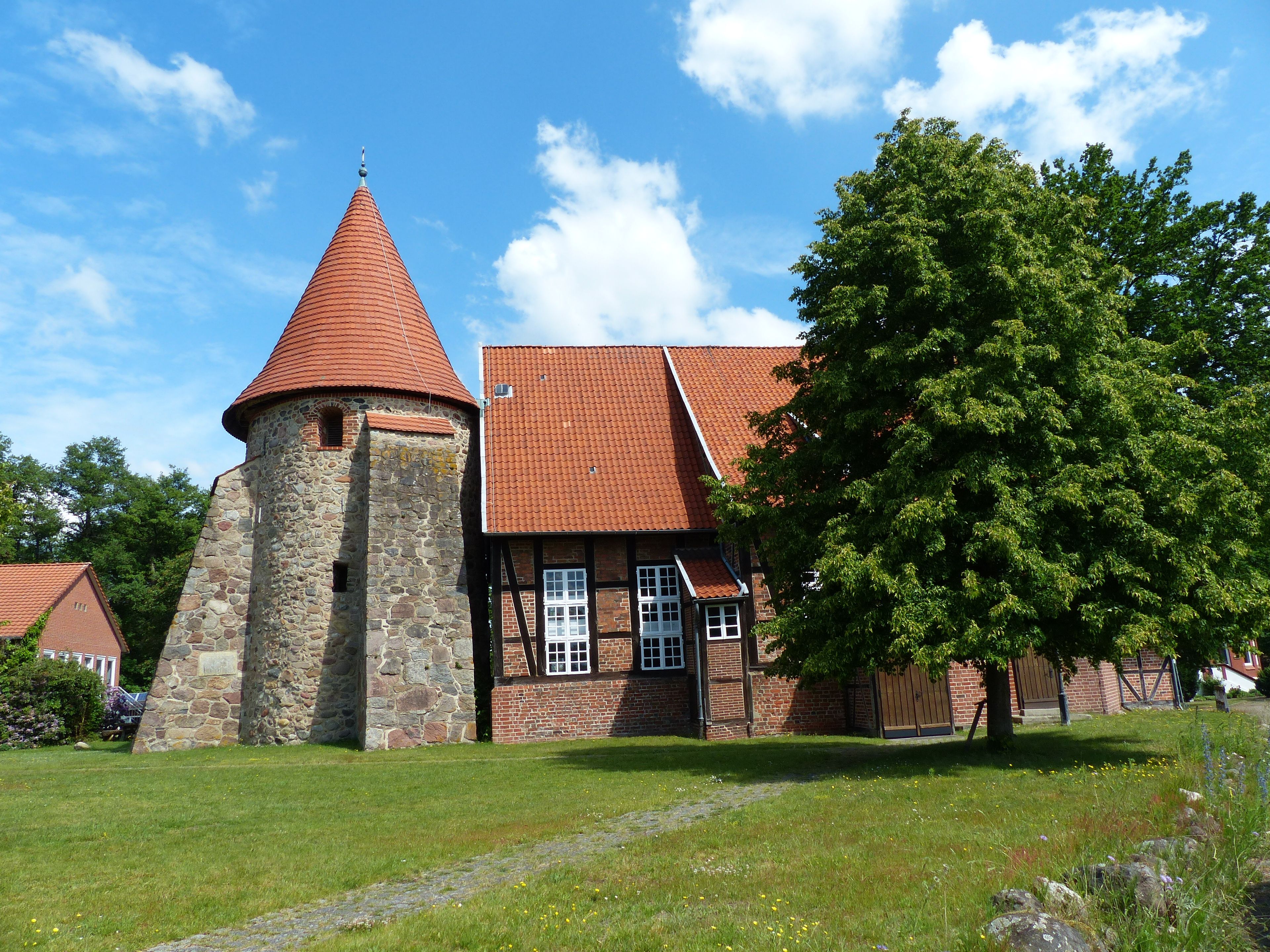 A historic building with a stone tower and red-tiled roof, surrounded by lush greenery.