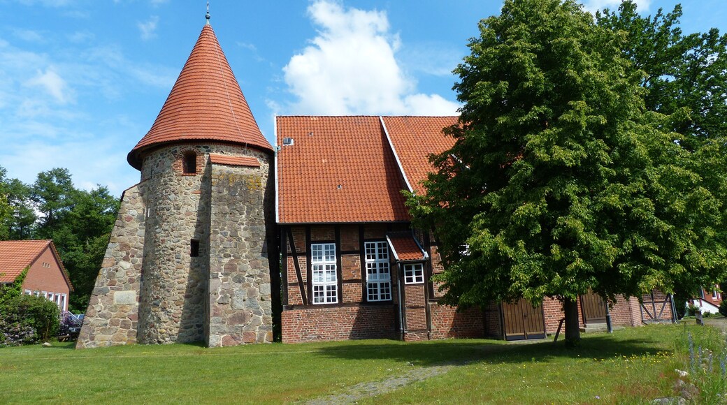A historic building with a stone tower and red-tiled roof, surrounded by lush greenery.