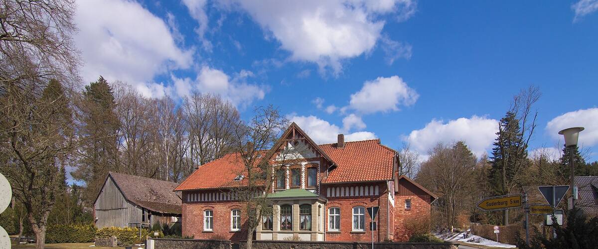 A large brick house with a red-tiled roof, surrounded by trees and a clear blue sky.