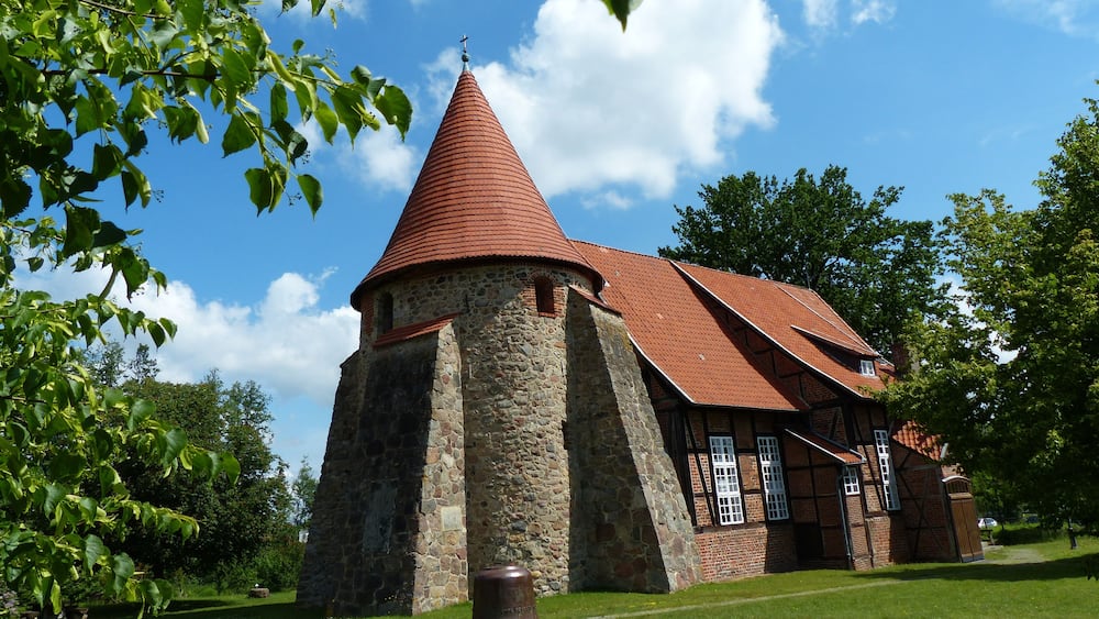 Eine Kirche mit einem spitzen Turmdach und Steinmauern, umgeben von viel Grün und blauem Himmel.