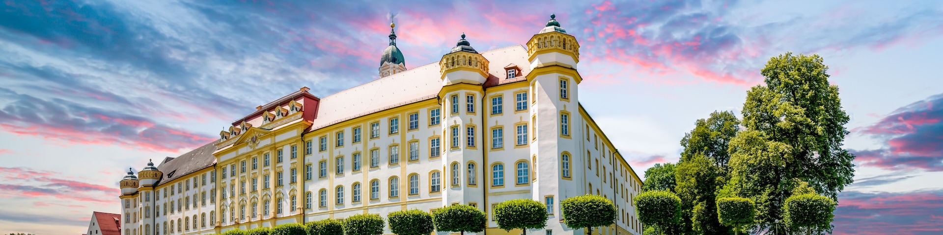 Un edificio grande y ornamentado, con múltiples torres y un techo rojo, rodeado de céspedes y árboles bien cuidados.
