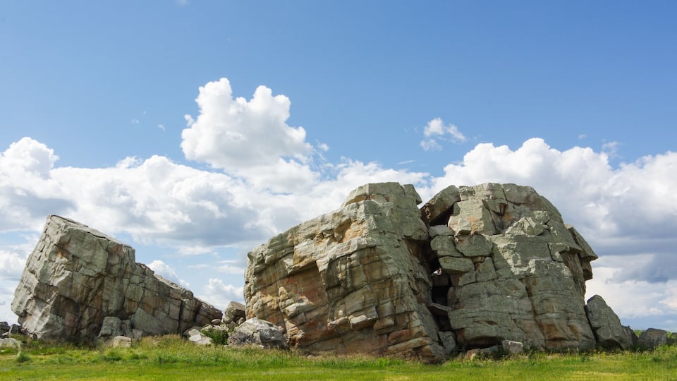 A large rock formation with a cave entrance, surrounded by a grassy field under a partly cloudy sky.