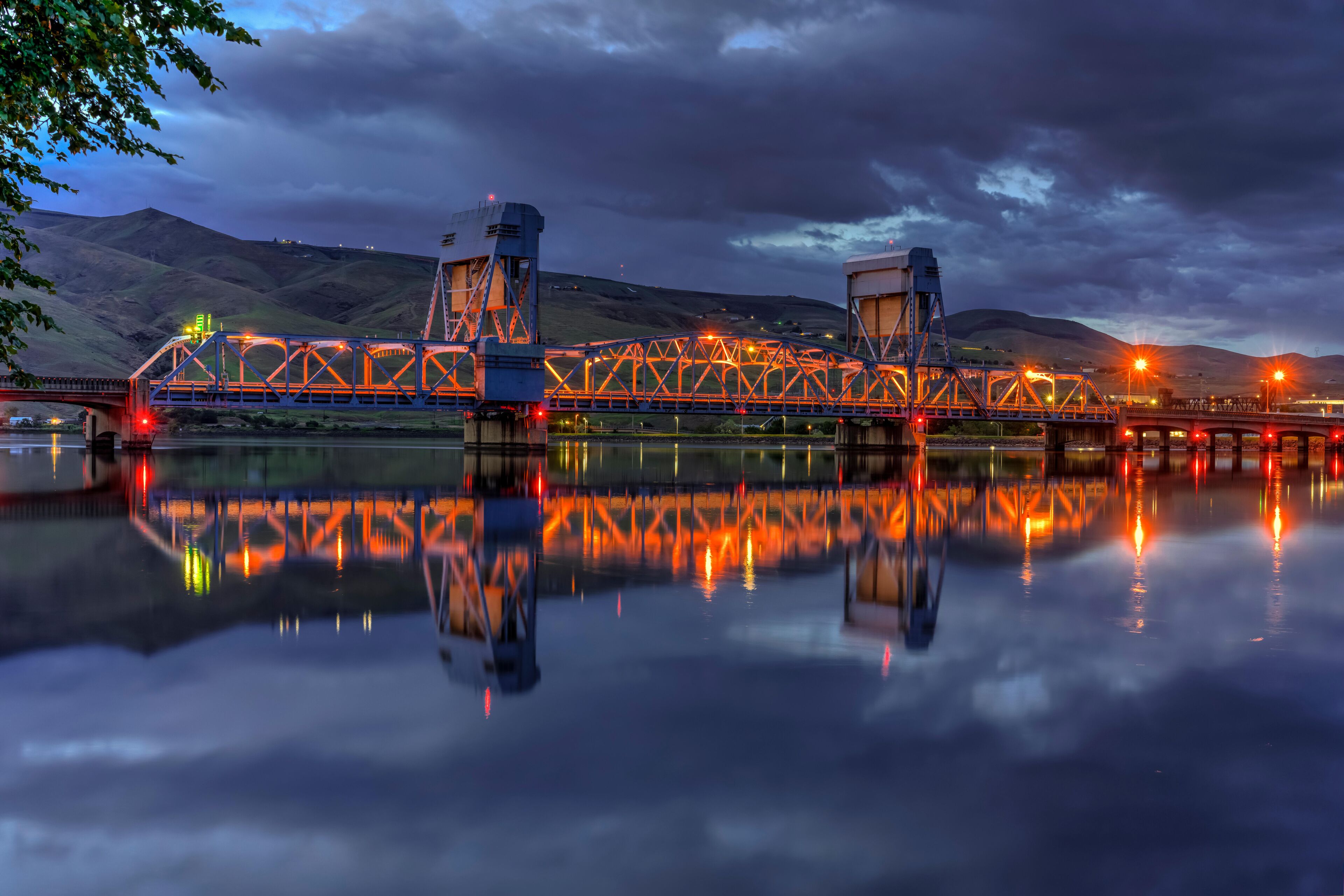 A bridge with a truss structure, reflecting on a calm body of water, with hills in the background.