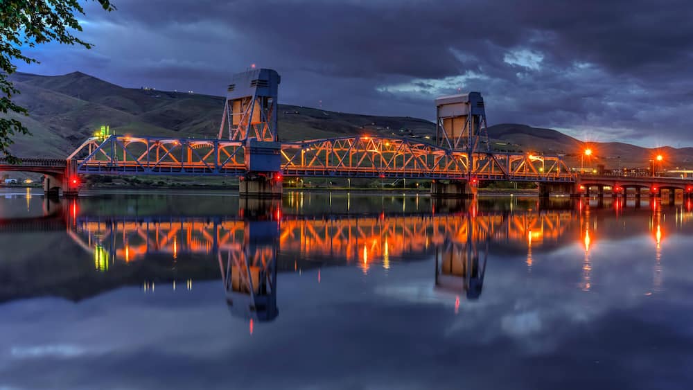 Eine Brücke mit Fachwerkkonstruktion spiegelt sich in ruhigem Wasser, im Hintergrund Hügel.