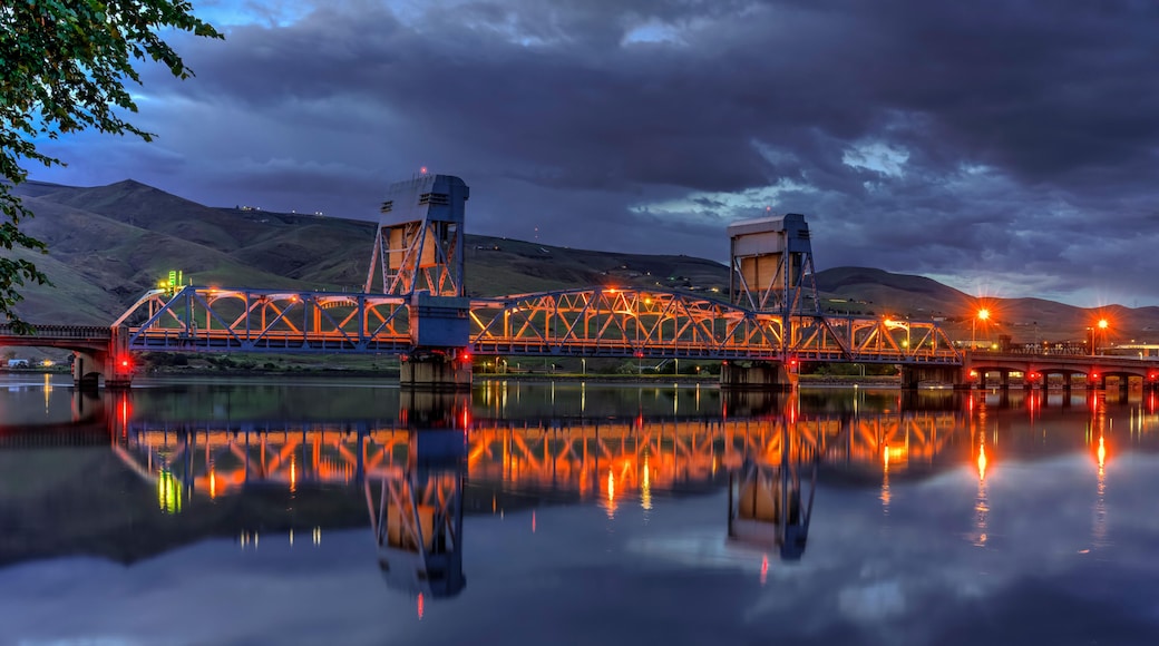 A bridge with a truss structure, reflecting on a calm body of water, with hills in the background.
