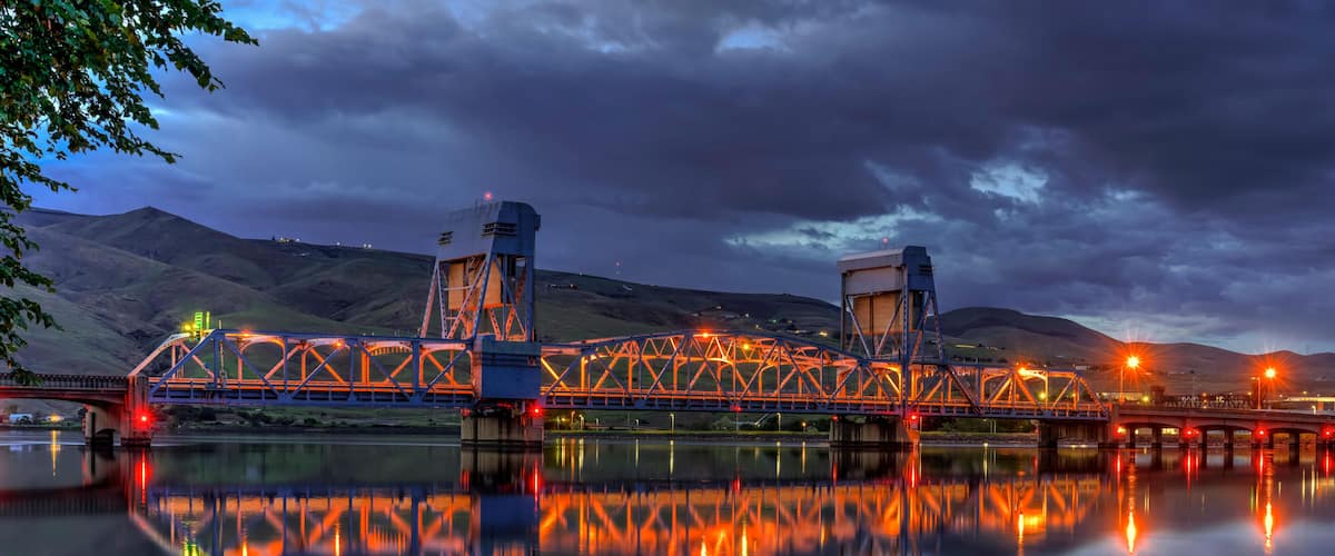 A bridge with a truss structure, reflecting on a calm body of water, with hills in the background.