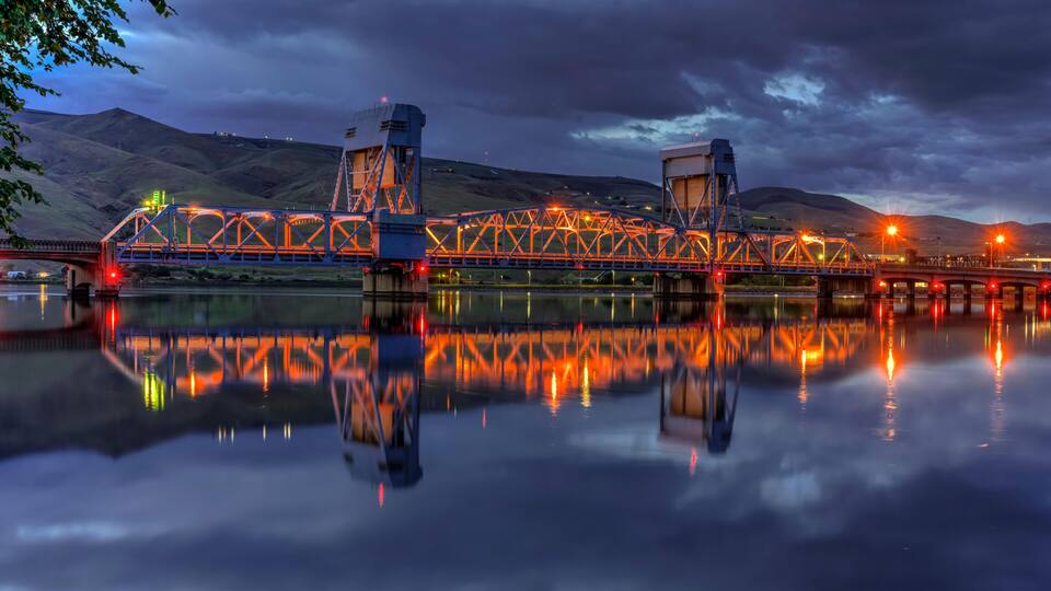 A bridge with a truss structure, reflecting on a calm body of water, with hills in the background.